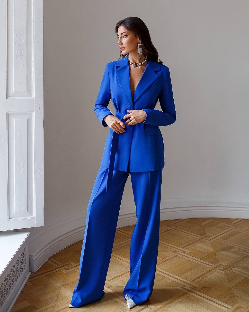 Woman wearing a blue suit standing in a room with wooden flooring and white walls.