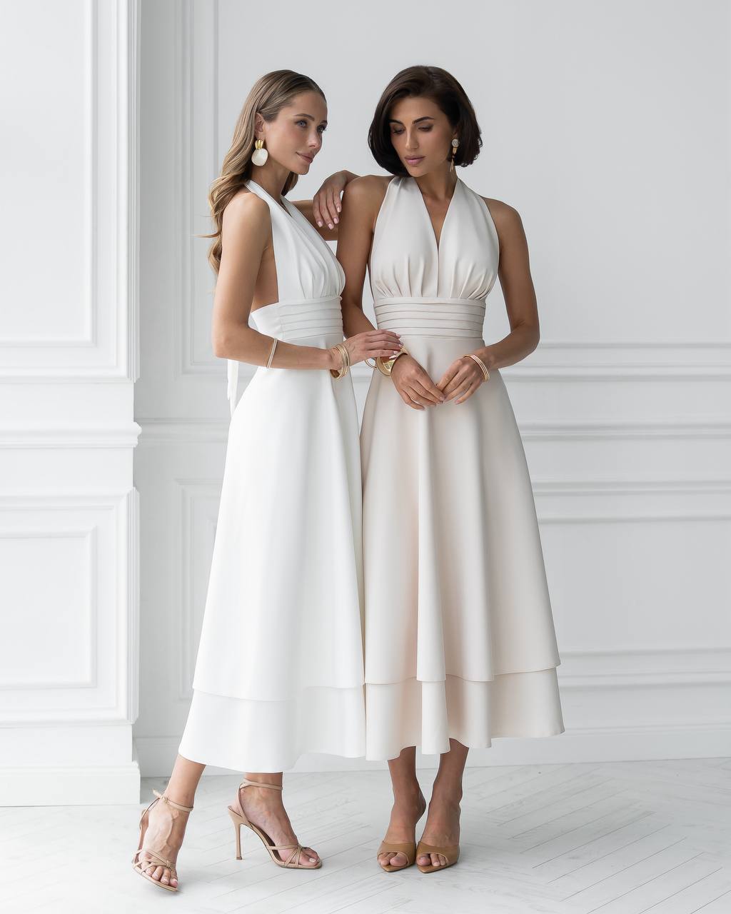 Two women wearing white halter neck dresses in a minimalistic setting.