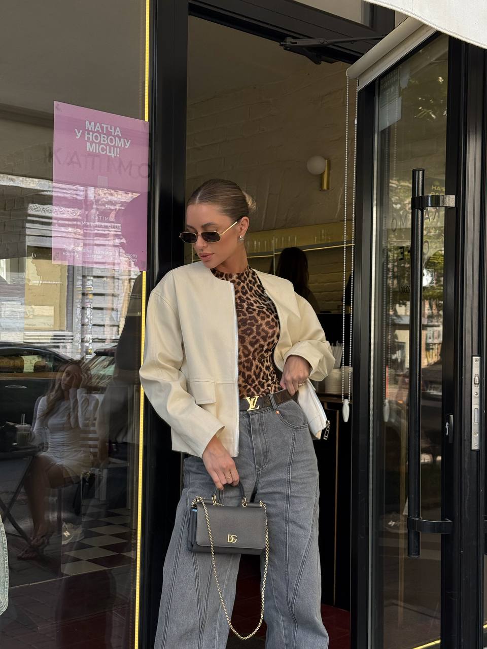 Woman exiting a building wearing sunglasses, a white jacket, leopard print top, and high-waisted jeans.