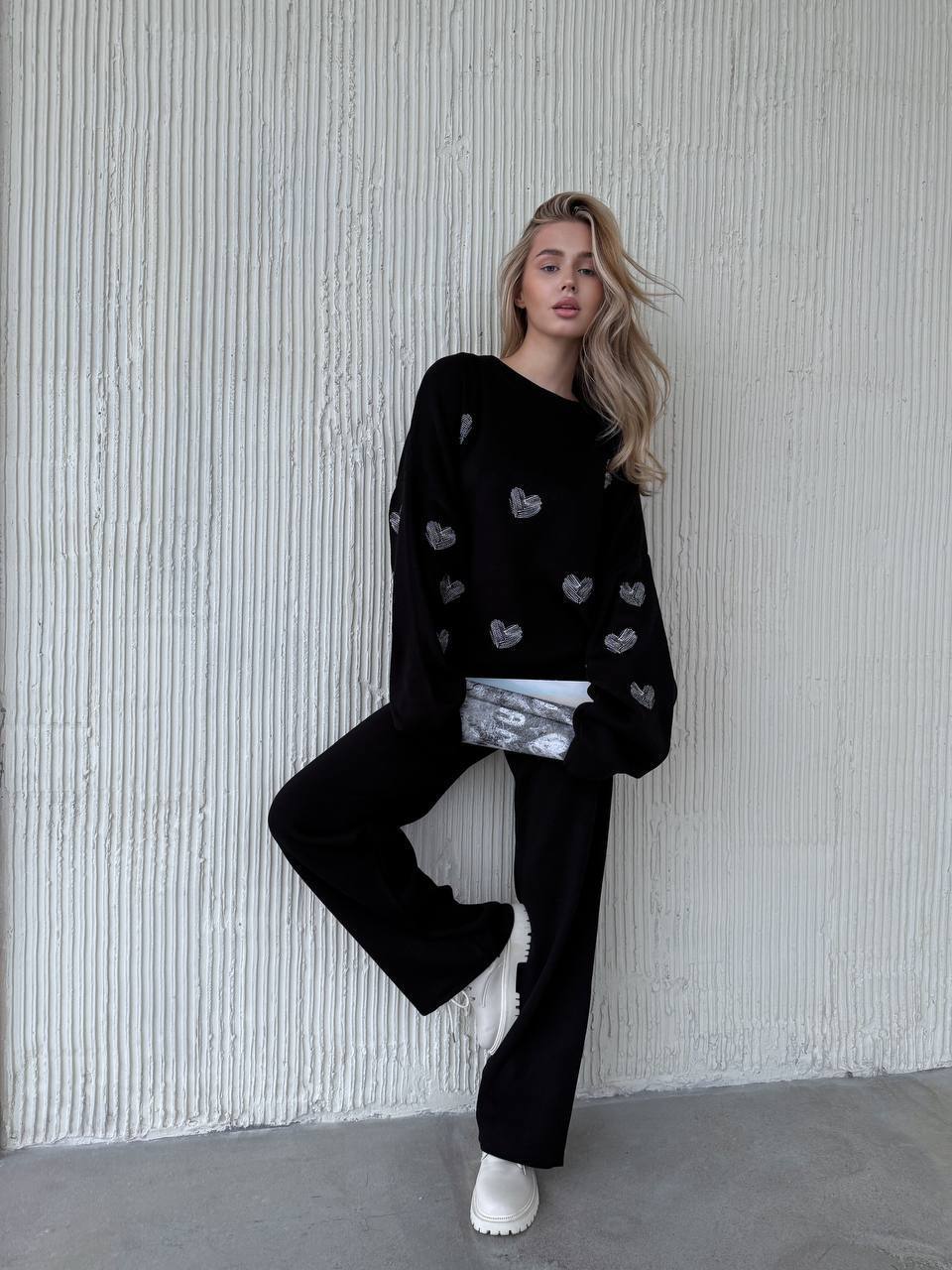 Person wearing a black sweater with heart patterns and black pants against a textured white wall.