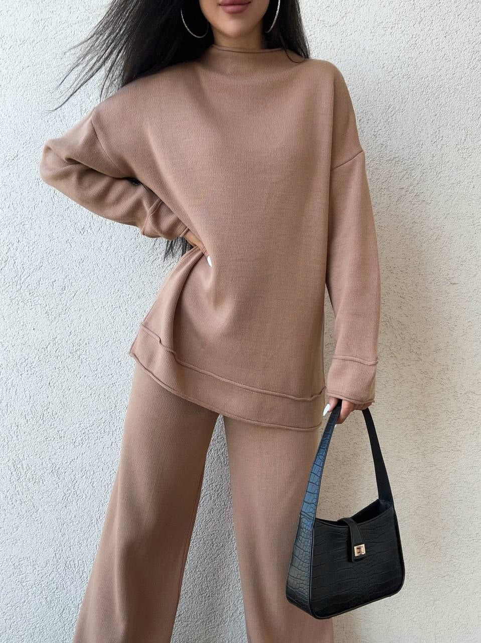 Person wearing a beige tracksuit holding a black handbag against a light background