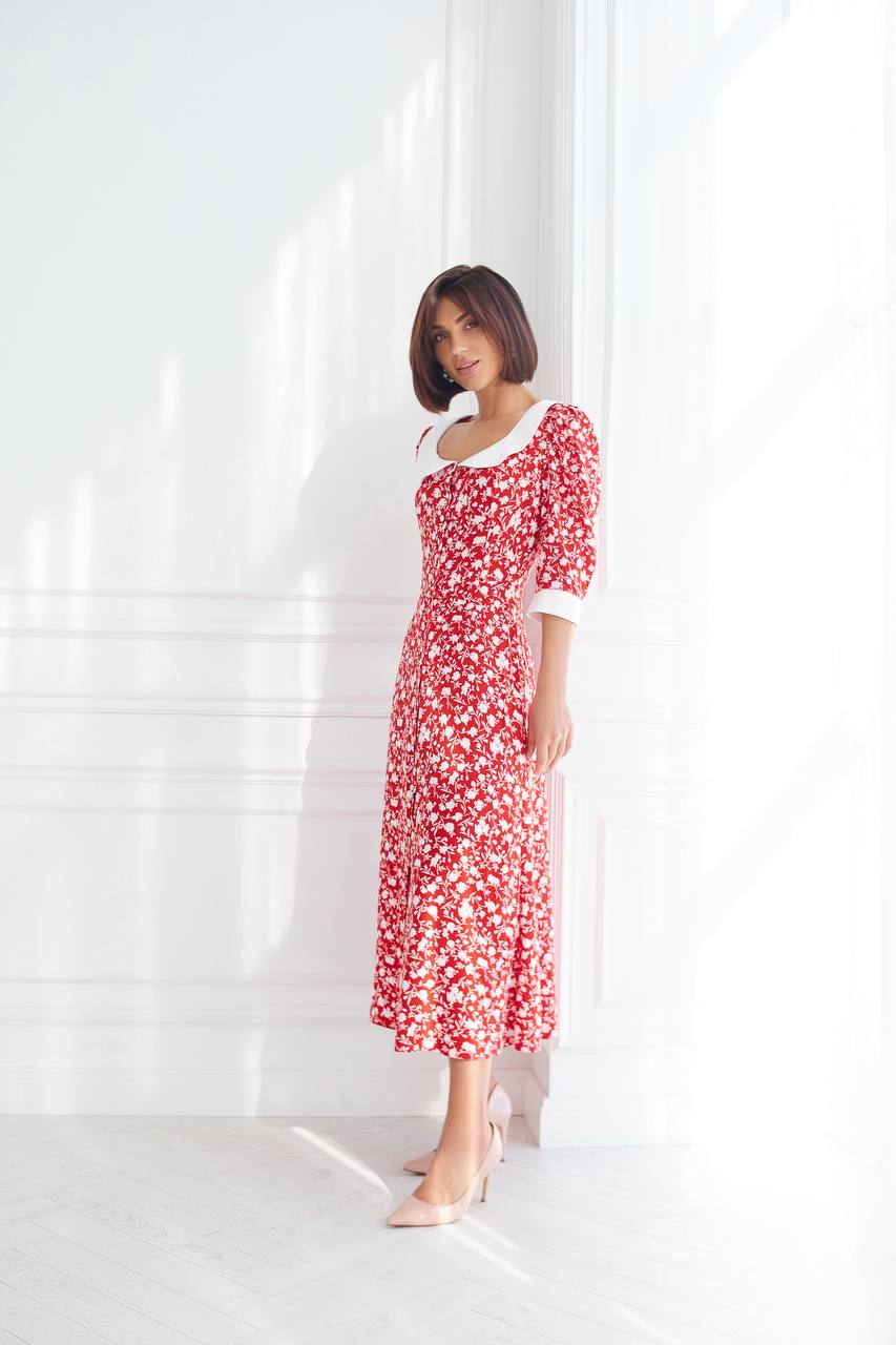 Woman wearing a red floral dress standing against a white curtain.