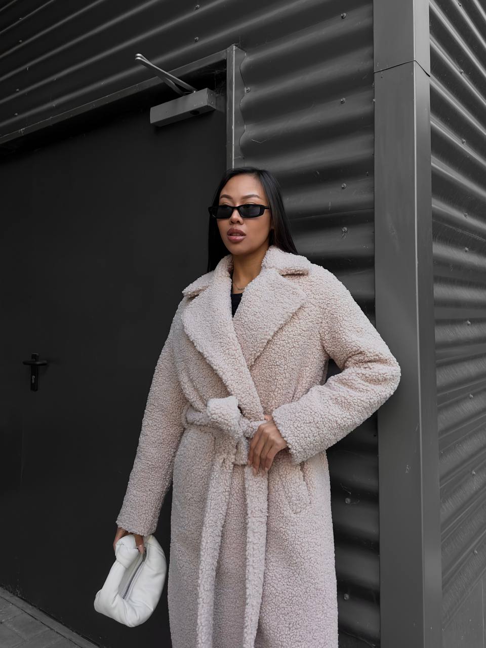 Woman wearing a beige teddy coat standing against a dark wall.