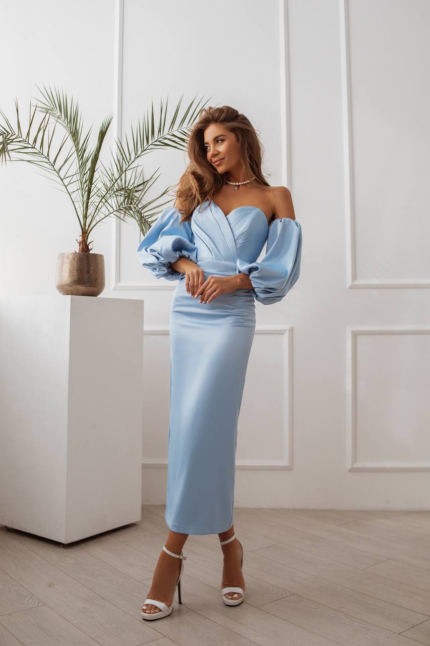 Woman in a light blue dress standing in a room with a plant and white wall.