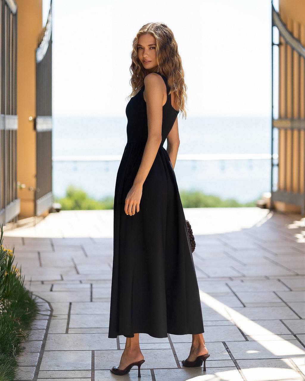 A woman wearing a sleeveless black maxi dress with a side cutout, standing on a stone walkway with a bright outdoor background.