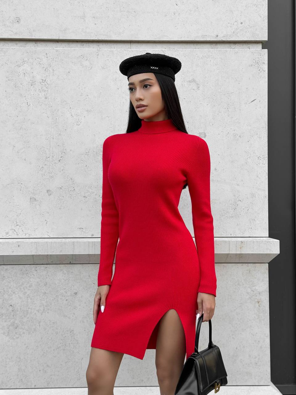 Woman wearing a red dress with a black beret and black handbag against a light gray wall.