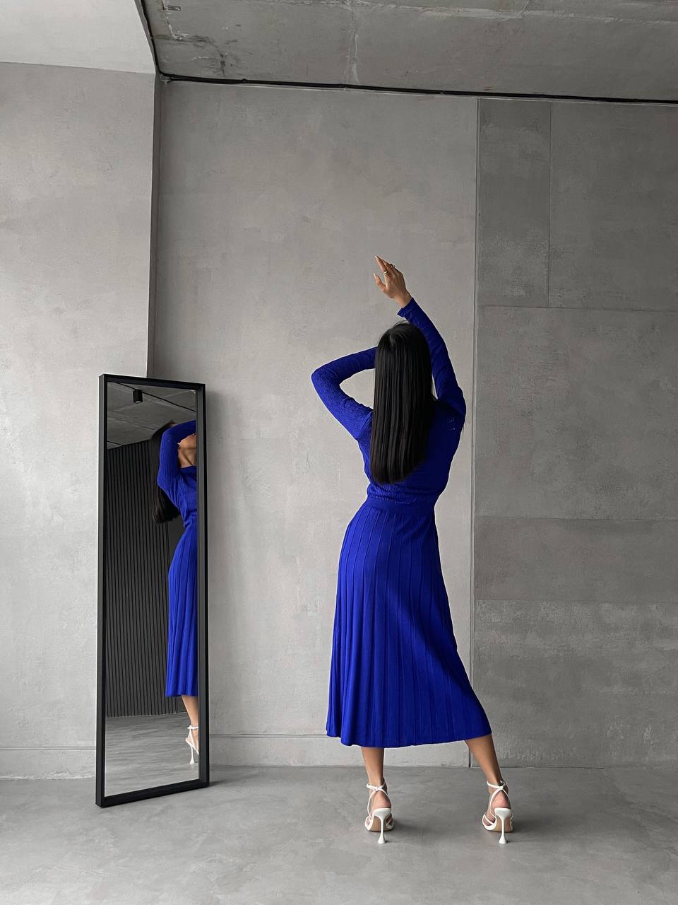 Woman in a blue dress standing in front of a mirror in a minimalistic room.