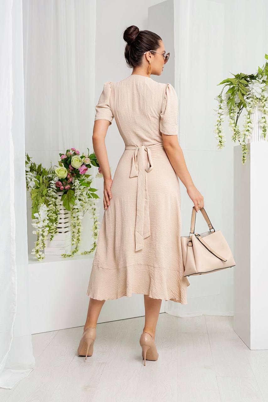 Woman in a beige dress holding a matching handbag in a minimalistic setting with plants.