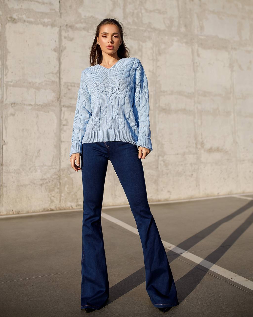Woman wearing a light blue cable knit sweater and dark blue flared jeans against a concrete wall.