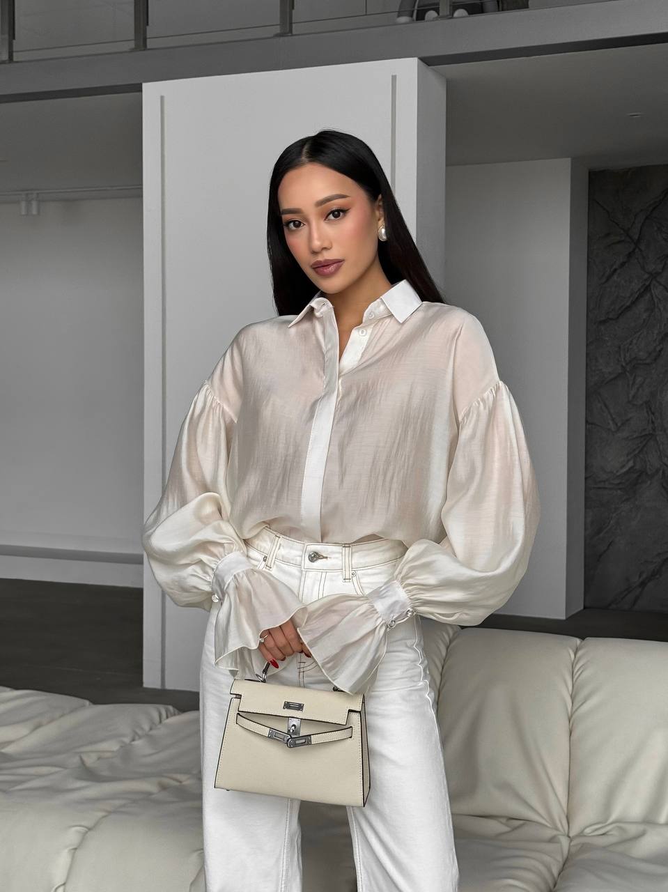 Woman in a white outfit holding a beige handbag in a minimalistic room.
