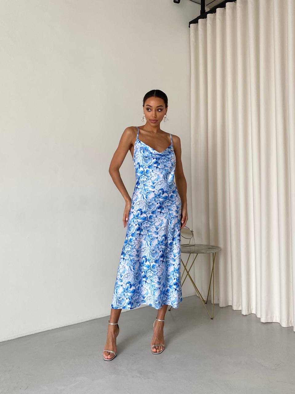 Woman wearing a blue floral dress standing in a minimalistic room with white curtains.