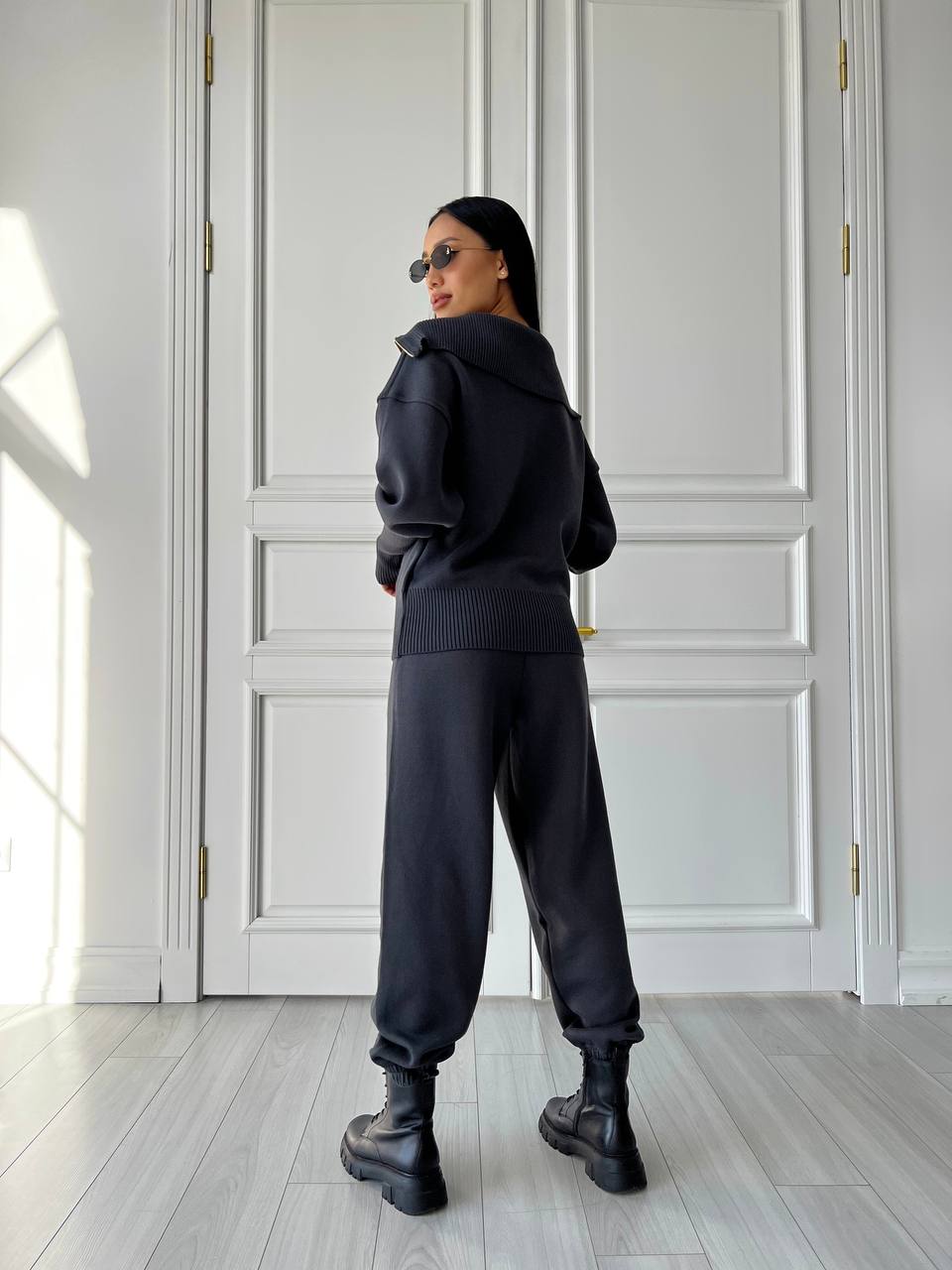 Person wearing a black hoodie and pants in a room with white walls and wooden flooring.