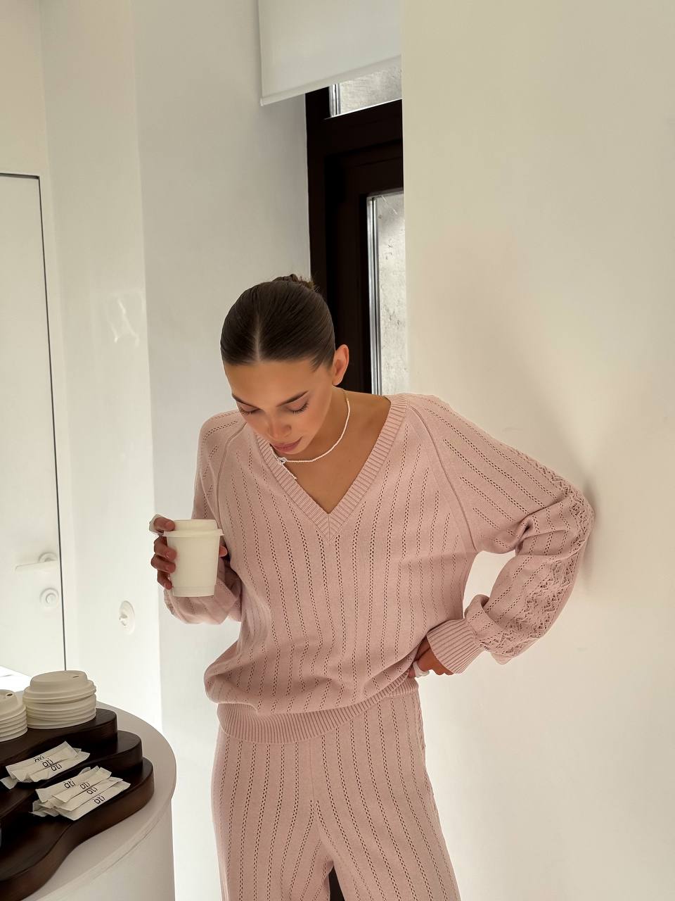 Woman in a pink knit outfit holding a cup in a minimalistic room.
