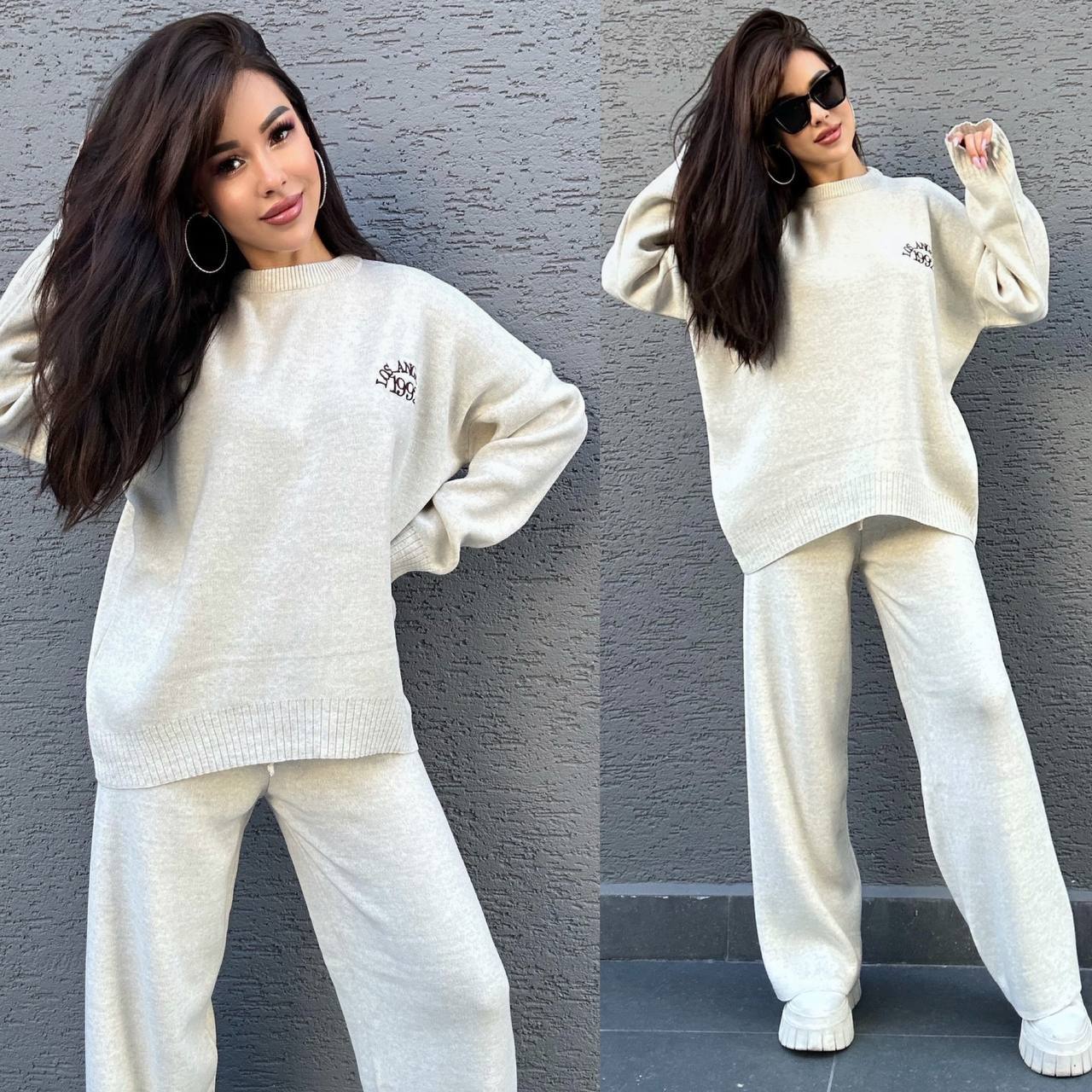 Woman wearing a white sweater and pants set against a gray wall.