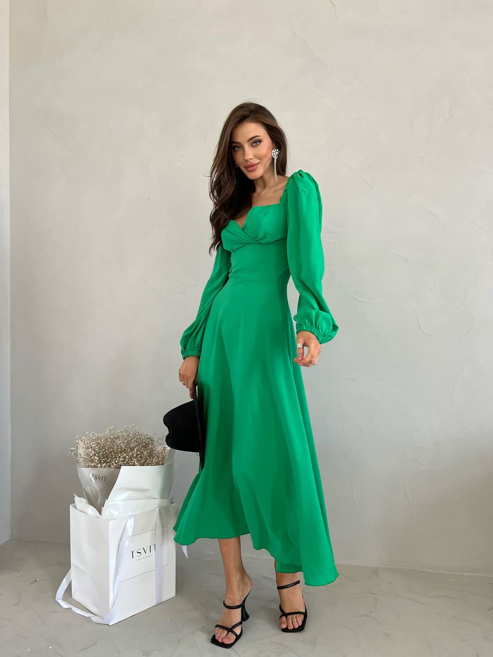 Woman wearing a green dress holding a black handbag against a plain background