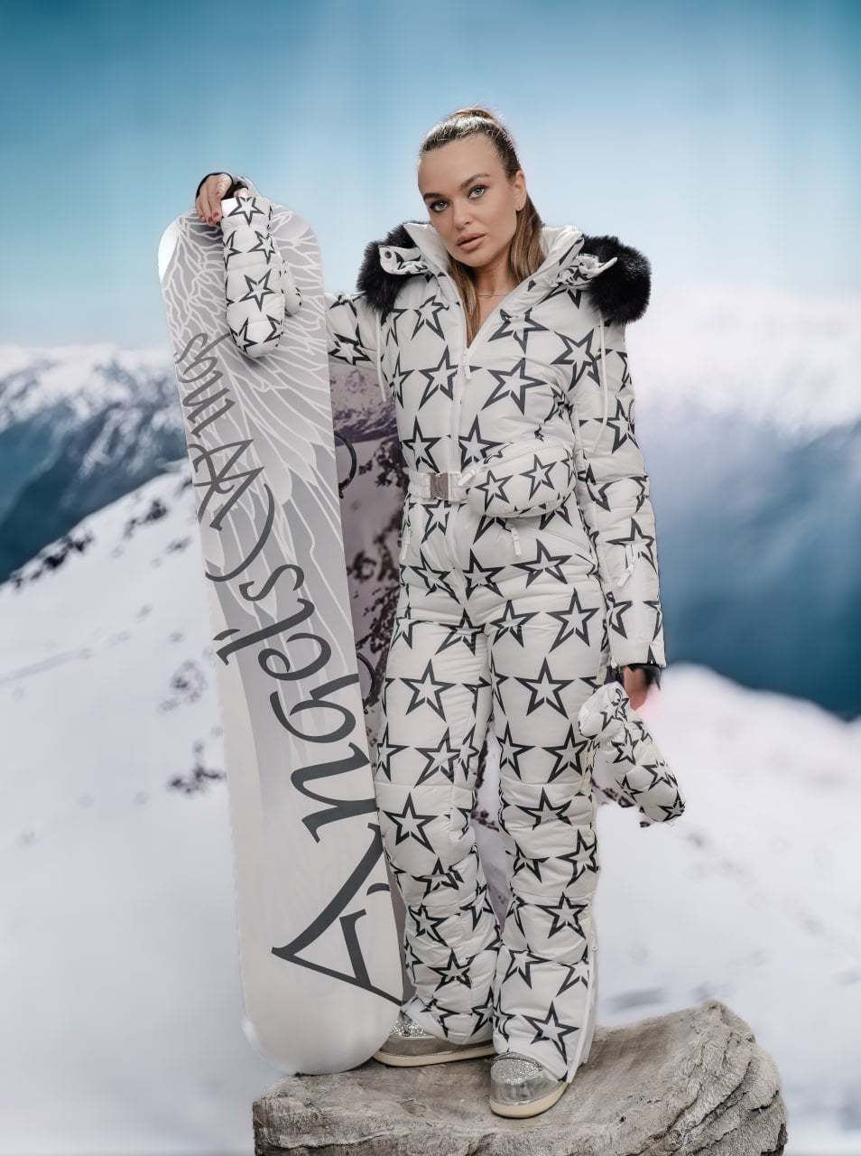 Person in star-patterned snowsuit holding a snowboard with mountain background