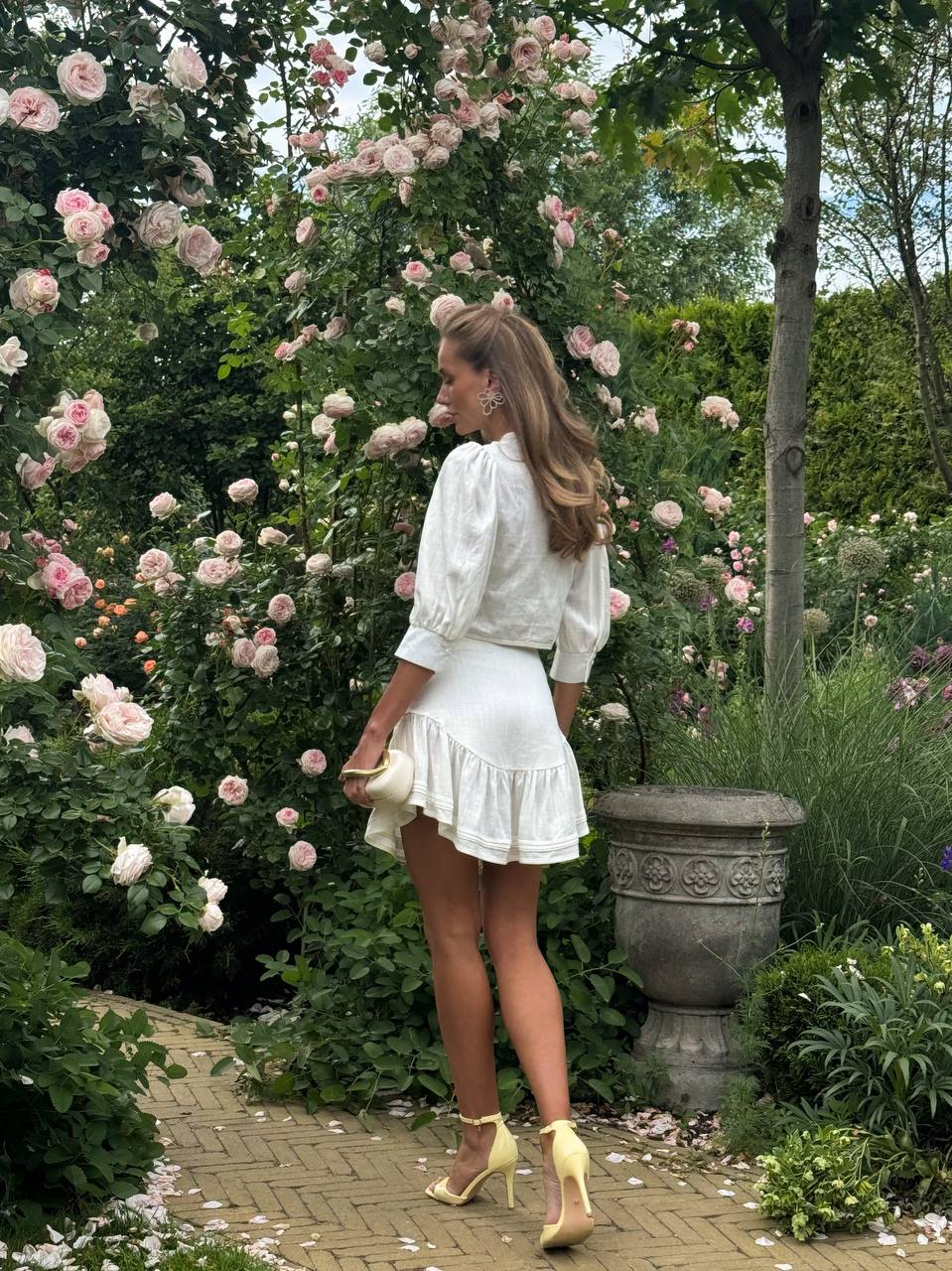 Woman in a white outfit standing in a garden with pink roses and greenery.