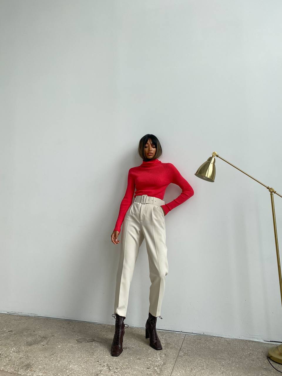 Person wearing a red sweater and white pants standing against a plain wall with a gold lamp.