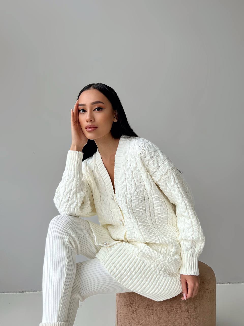 Woman wearing a white knit outfit sitting on a stool against a plain background