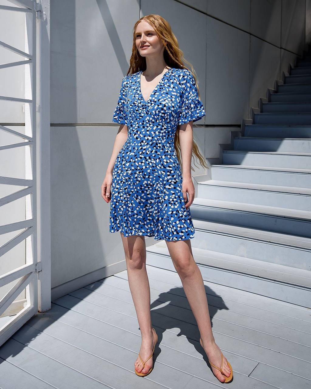 Woman in a blue floral dress standing on a sunlit outdoor staircase.