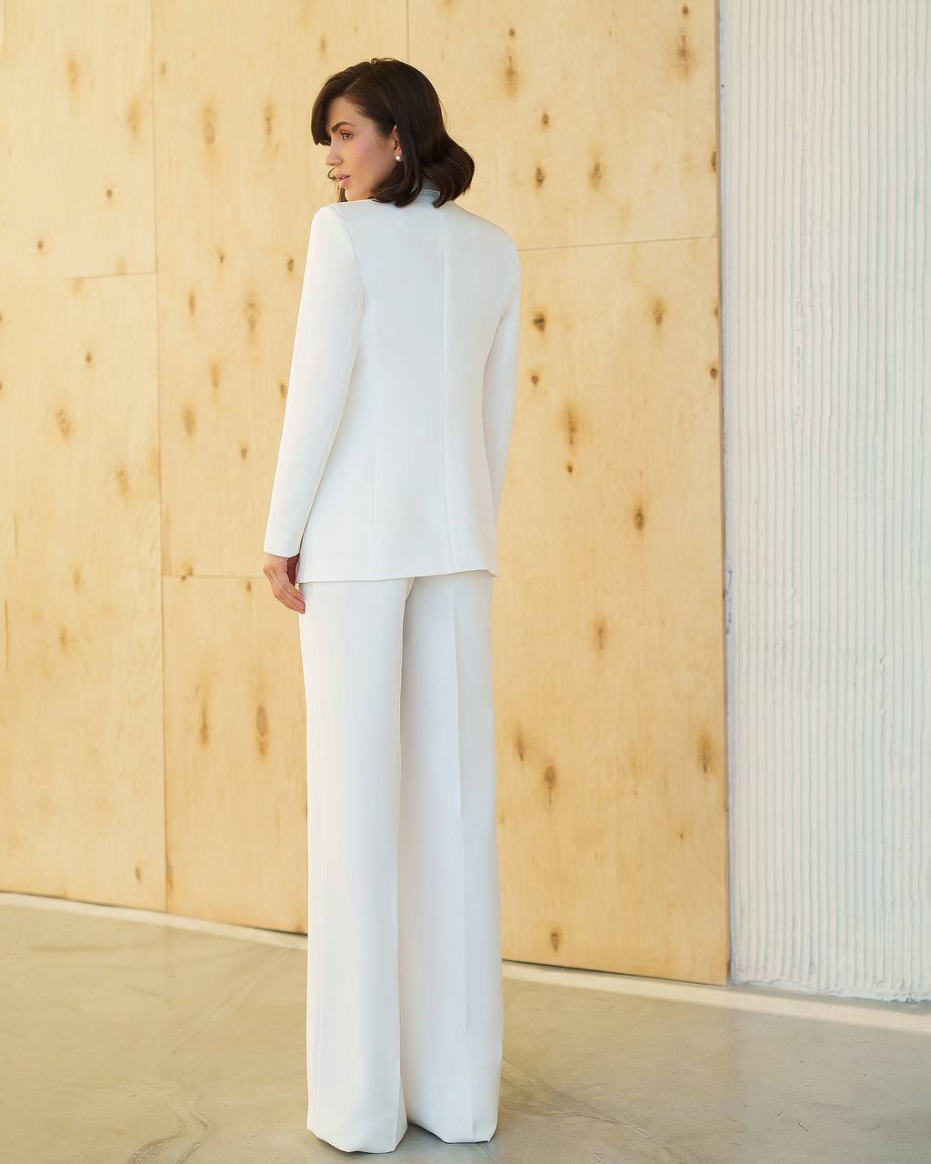 Woman wearing a white suit standing against a wooden wall.
