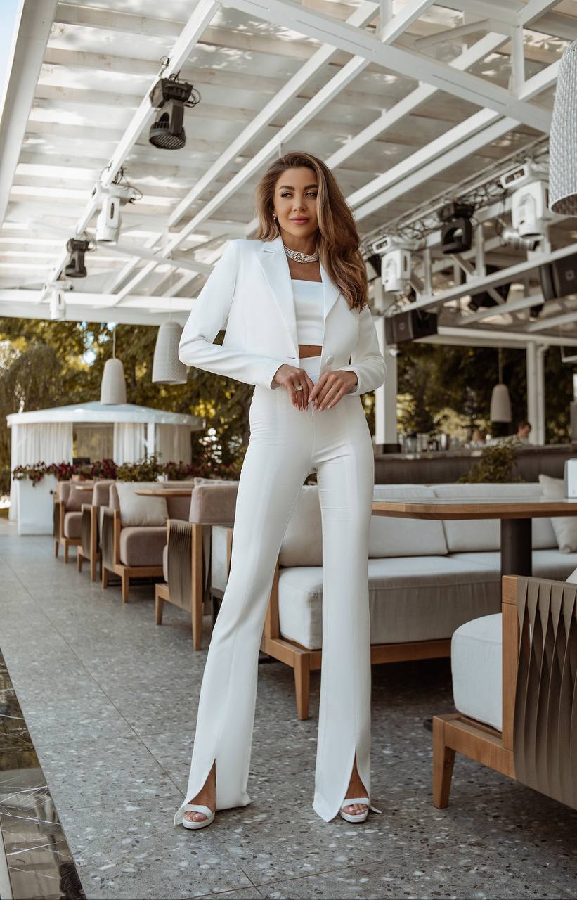 Woman in a white suit standing in an outdoor setting with tables and chairs.