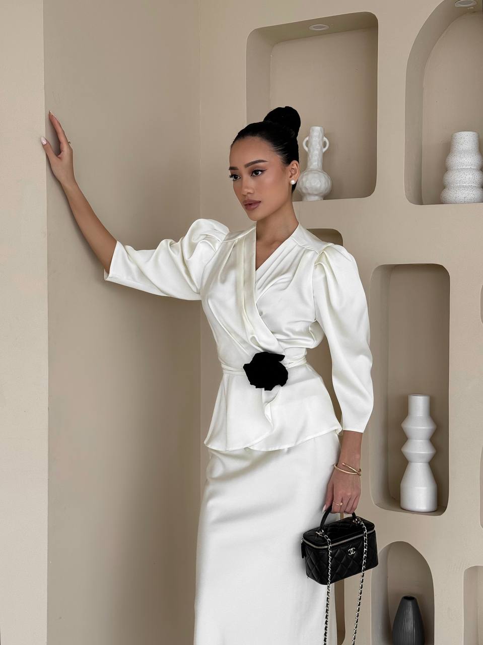 Woman in a white outfit with a black belt standing against a beige wall with decorative shelves.