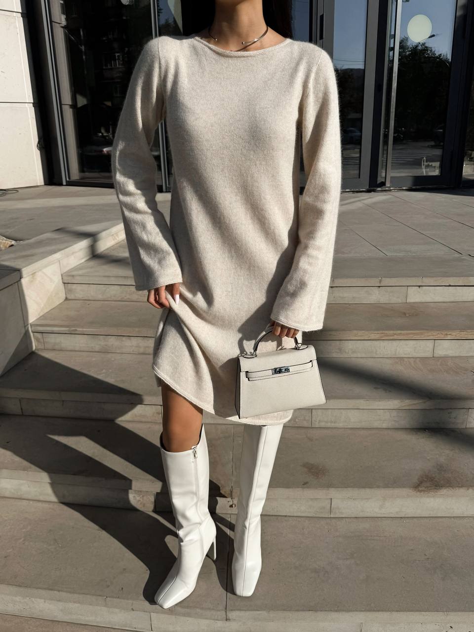 Person wearing a beige sweater dress and white boots, holding a matching handbag on steps.