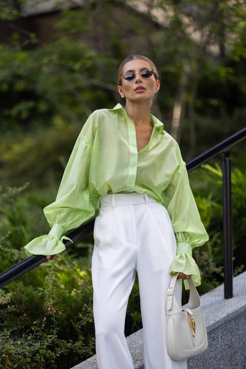 Woman wearing a light green blouse and white pants, standing outdoors with greenery in the background.