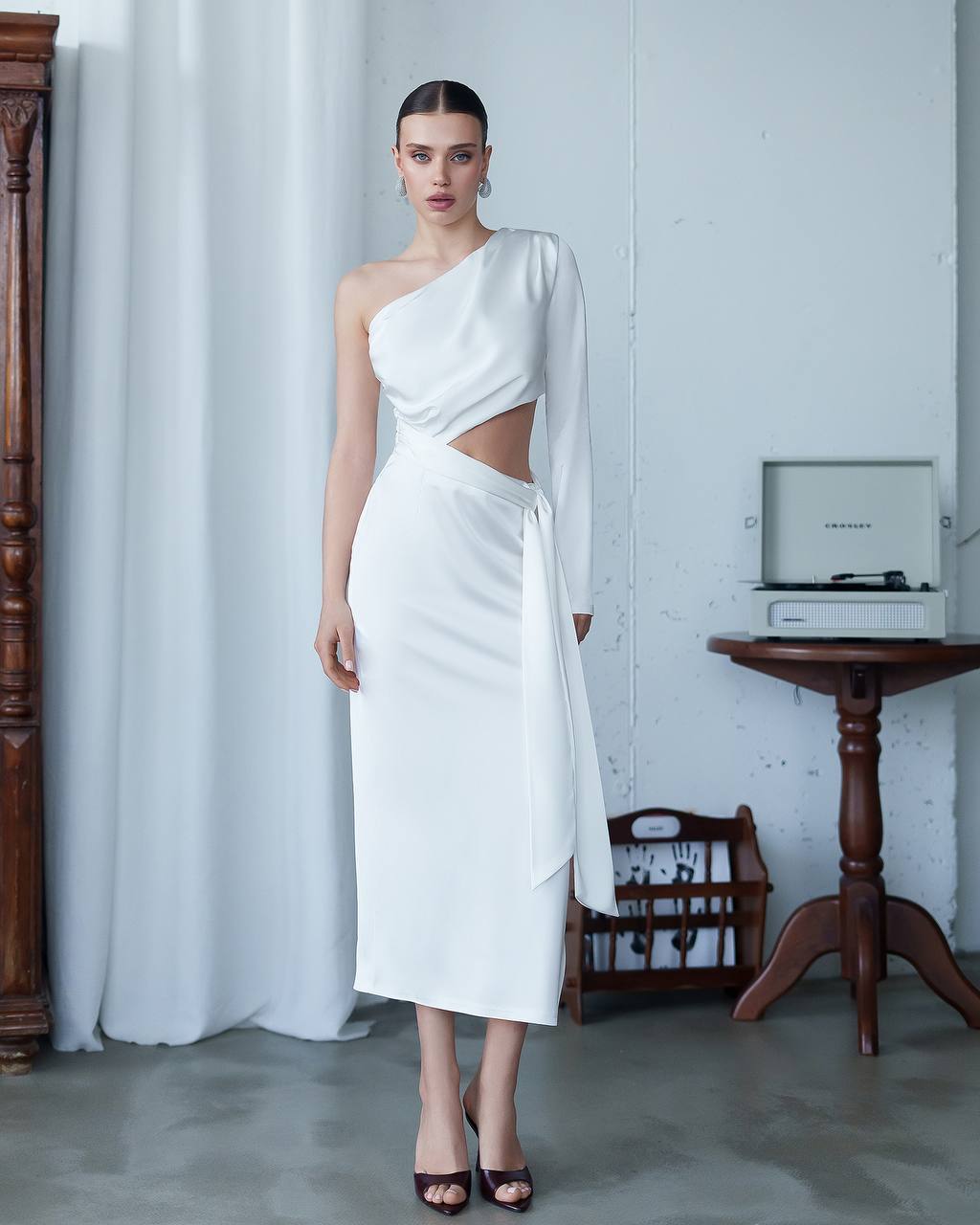 Woman wearing a white one-shoulder dress with cut-out design in a minimalistic room.