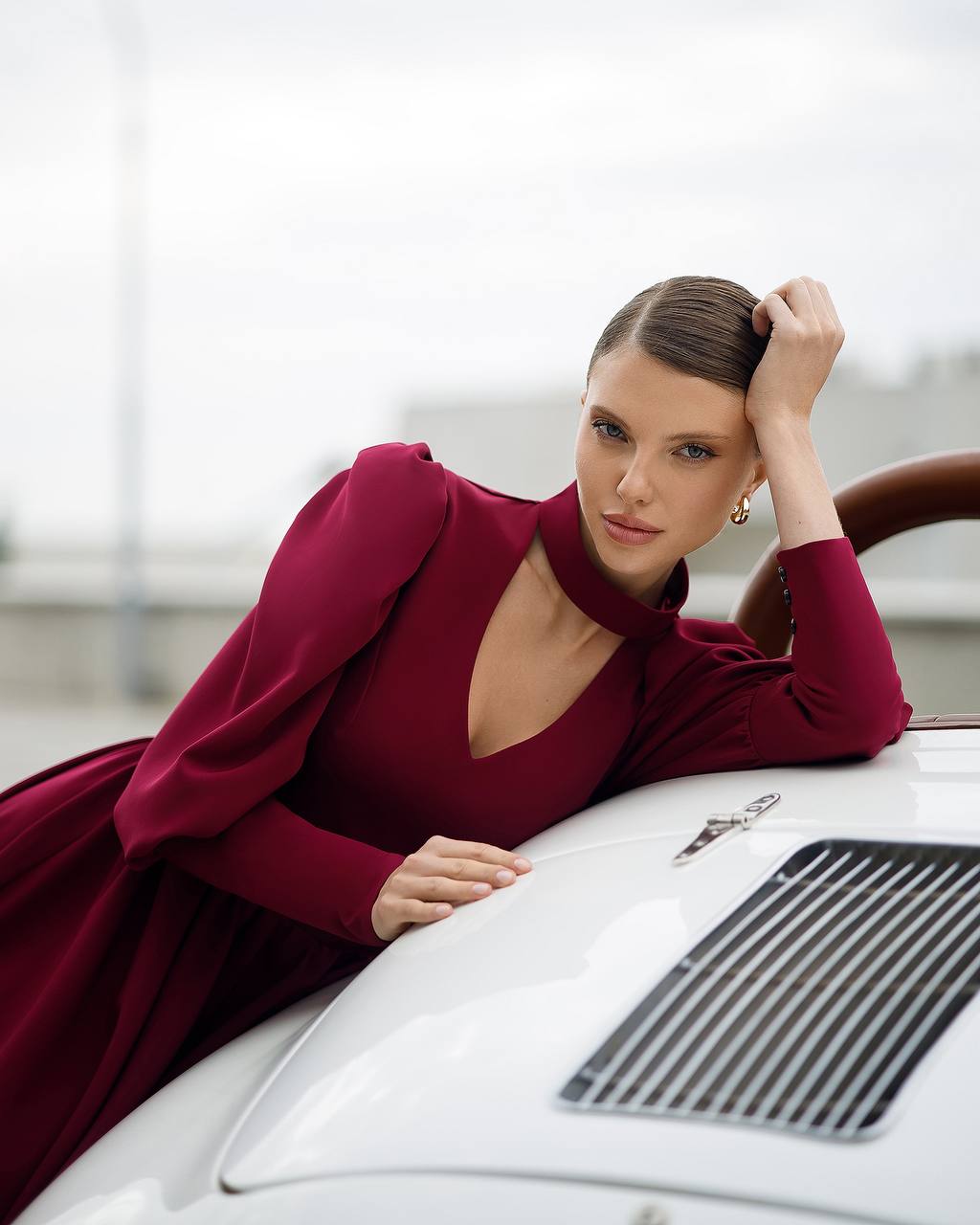 Woman in a burgundy dress leaning on a white car with a blurred background