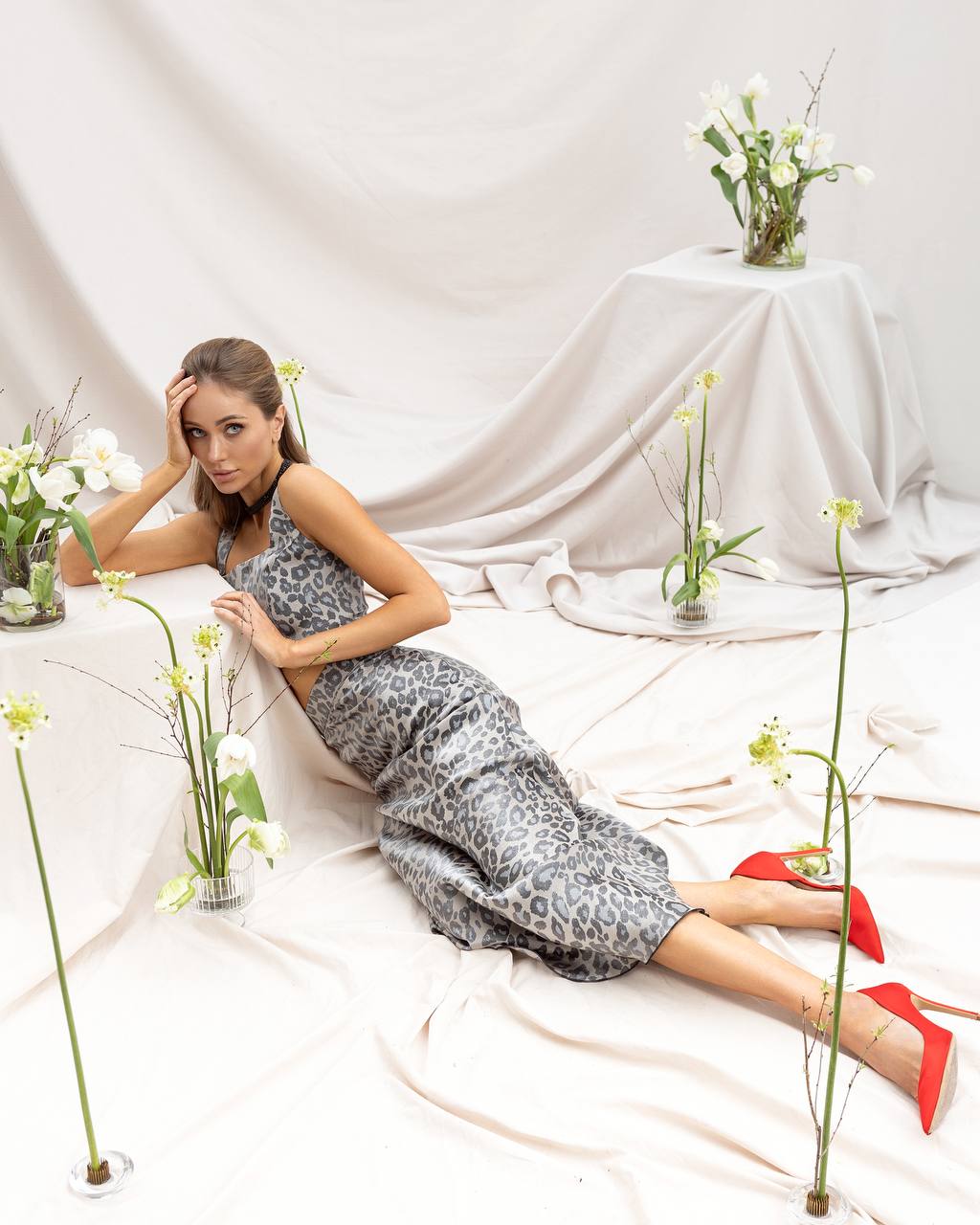 Woman in a patterned dress lying on a white surface with floral arrangements around her.