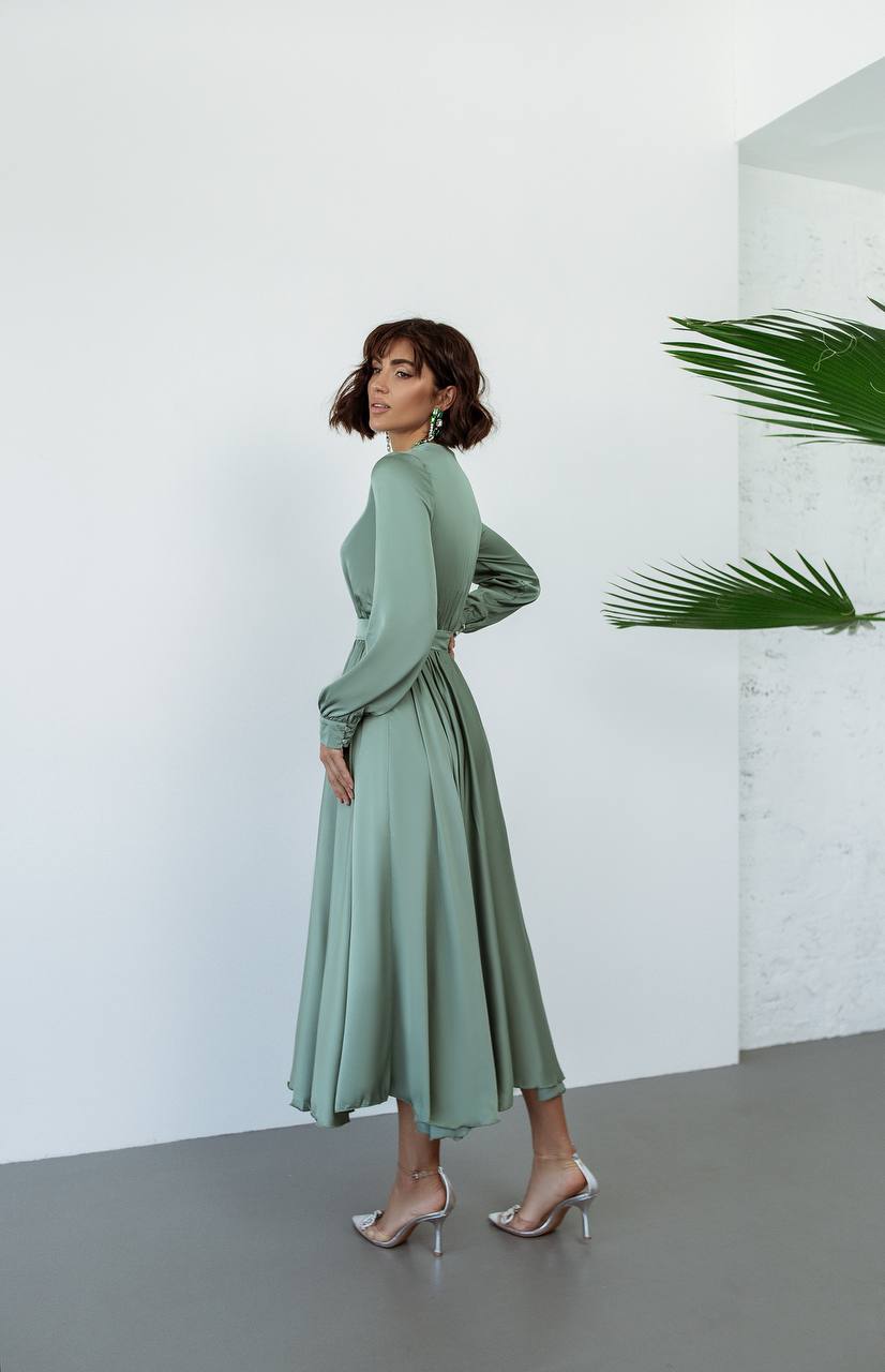 Woman wearing a green dress standing against a white wall with a plant in the corner.