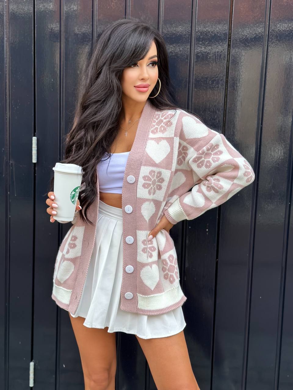 Woman wearing a patterned cardigan and white skirt, holding a Starbucks cup against a black background.