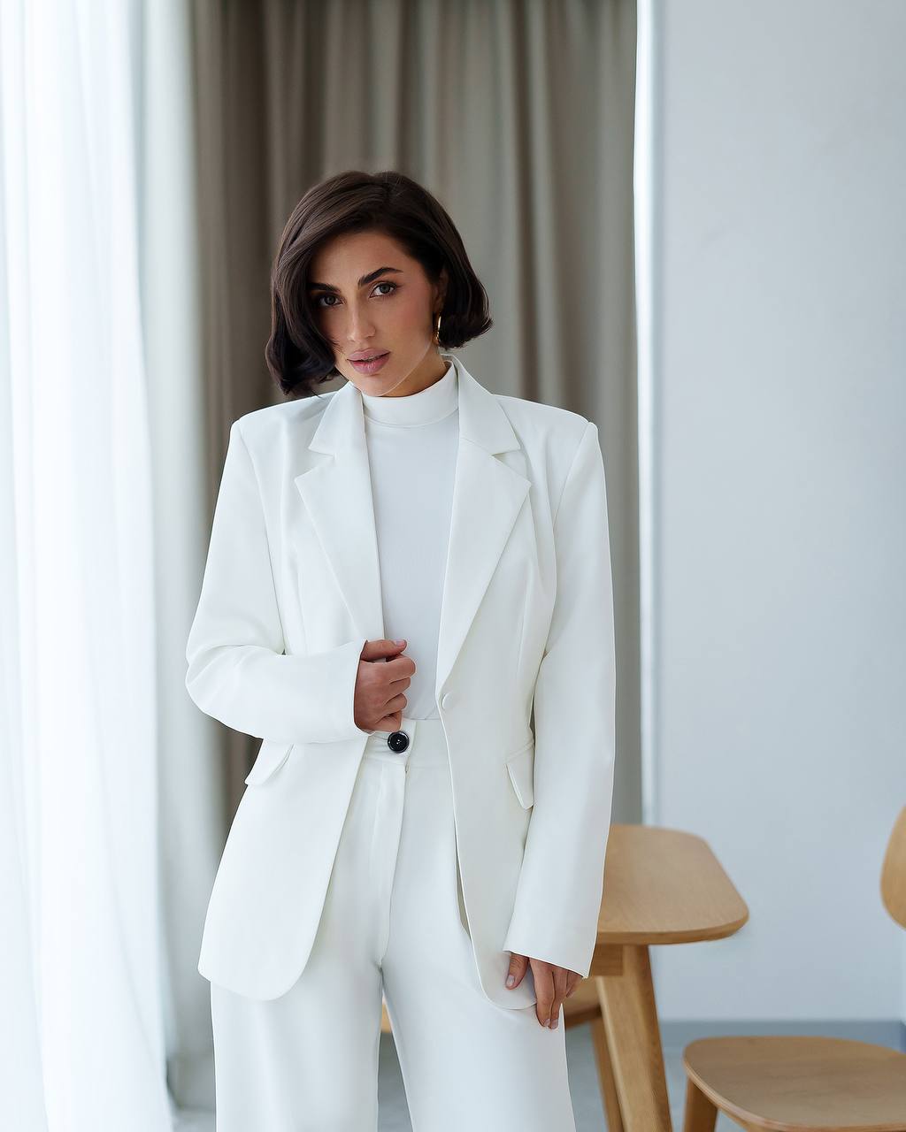 Woman wearing a white suit standing in a room with light-colored curtains and wooden furniture.