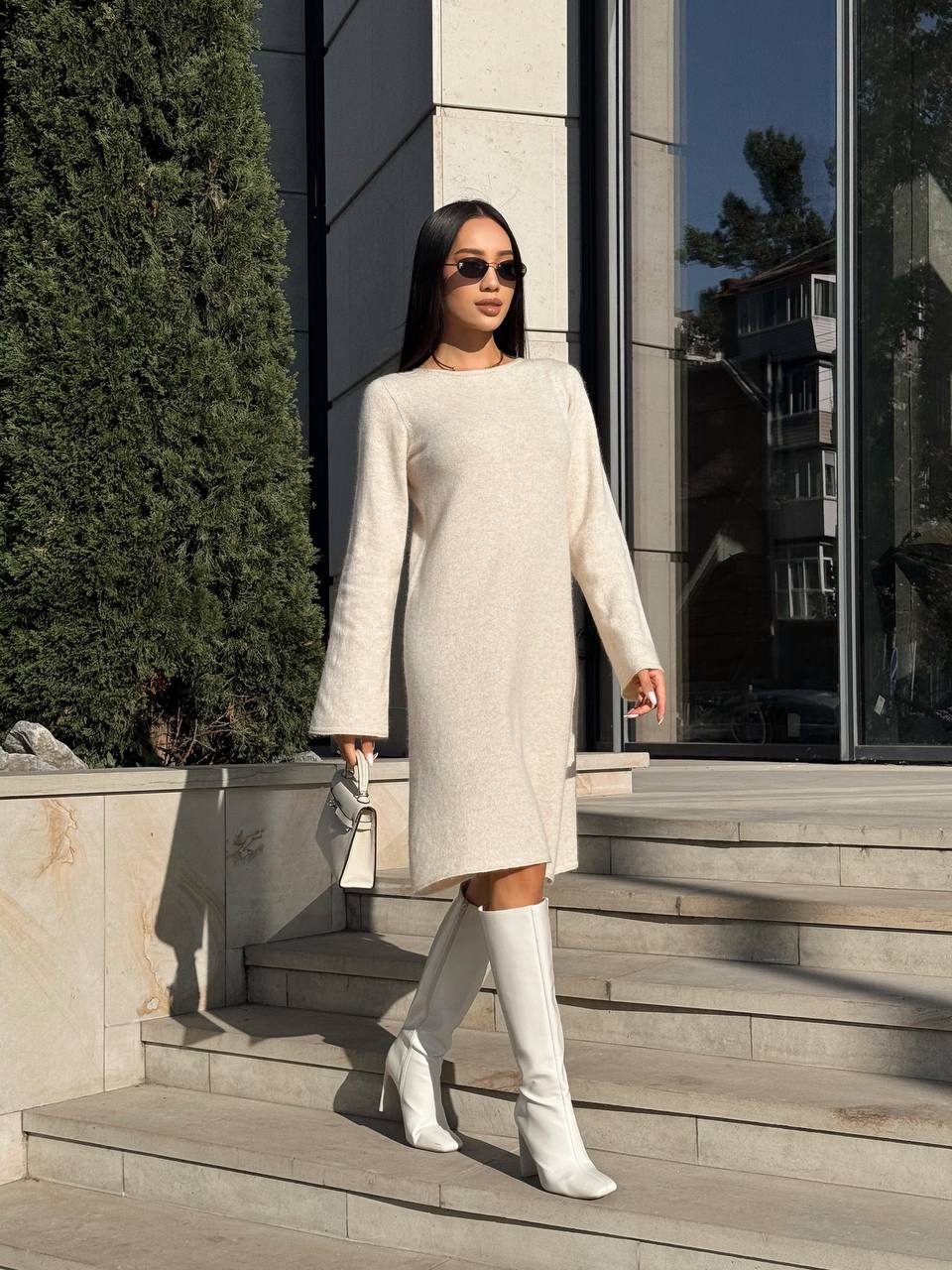 Woman in a light gray dress and white knee-high boots walking outside a building.