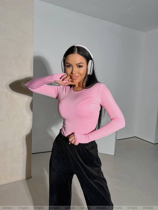 Person wearing a pink long-sleeve top and black pants in a minimalistic room.
