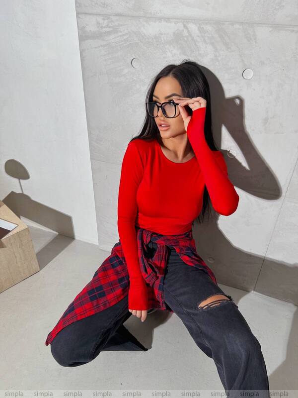 Woman in a red top and plaid shirt sitting against a light gray wall.
