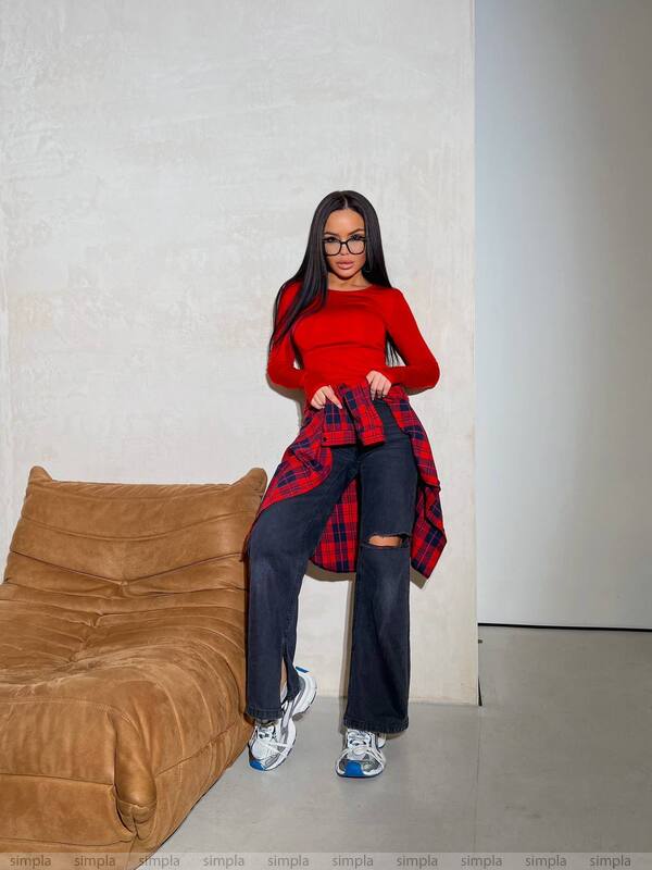 Person wearing a red plaid shirt over a red top with dark jeans, standing in a minimalistic room.