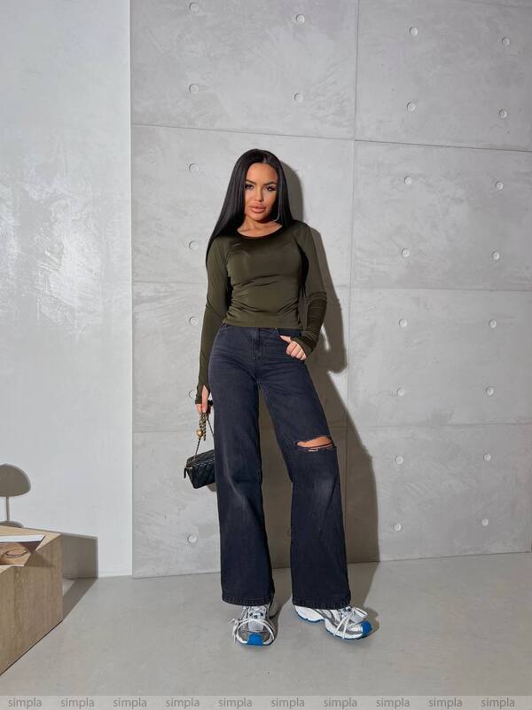 Woman wearing a green top and dark jeans standing against a gray wall.
