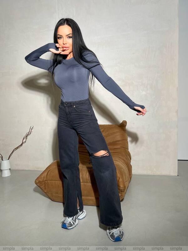 Woman in gray long-sleeve top and black jeans posing against a neutral wall.
