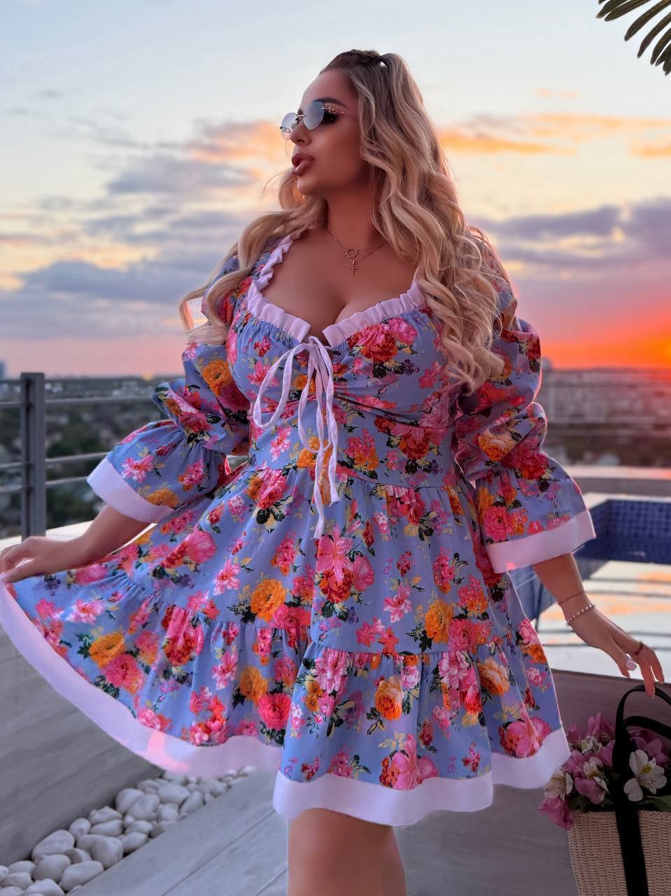 A woman wearing a light blue floral mini dress with pink and orange flowers, white ruffle trim, and a white drawstring tie, posing on a balcony at sunset.