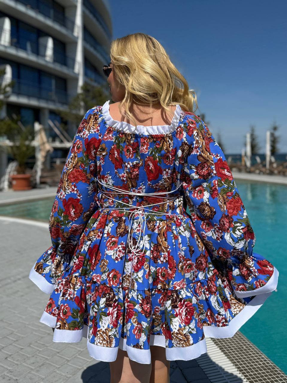 A woman stands with her back to the camera wearing a blue floral dress with white ruffled trim, photographed outdoors beside a swimming pool and modern building.