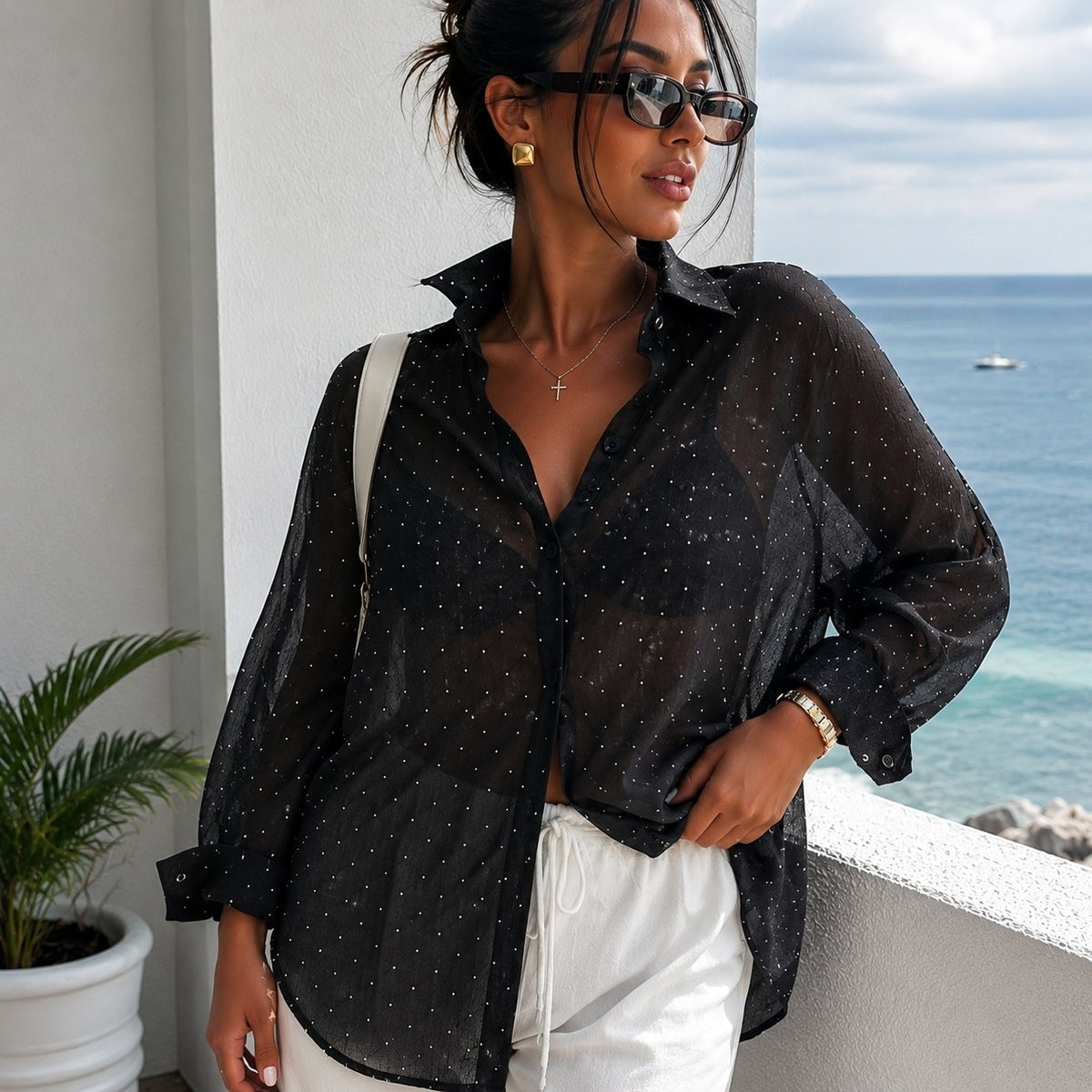 A woman wearing a sheer black button-up shirt with tiny white polka dots and white pants, standing on a balcony with the ocean in the background.