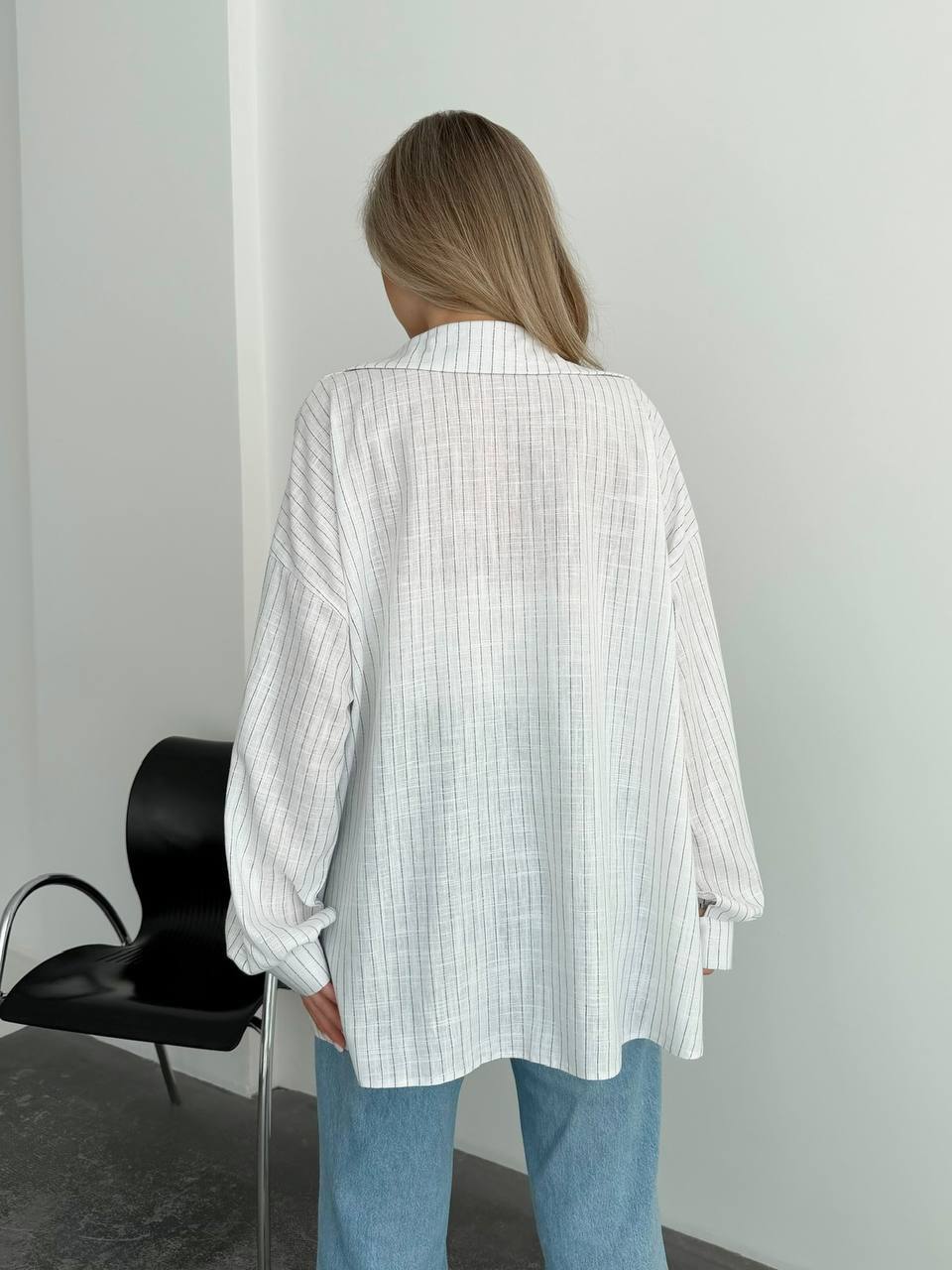 A woman viewed from behind wearing an oversized white button-up shirt with a subtle vertical stripe pattern and light blue jeans, standing in a minimalist room with a black chair against a white wall.