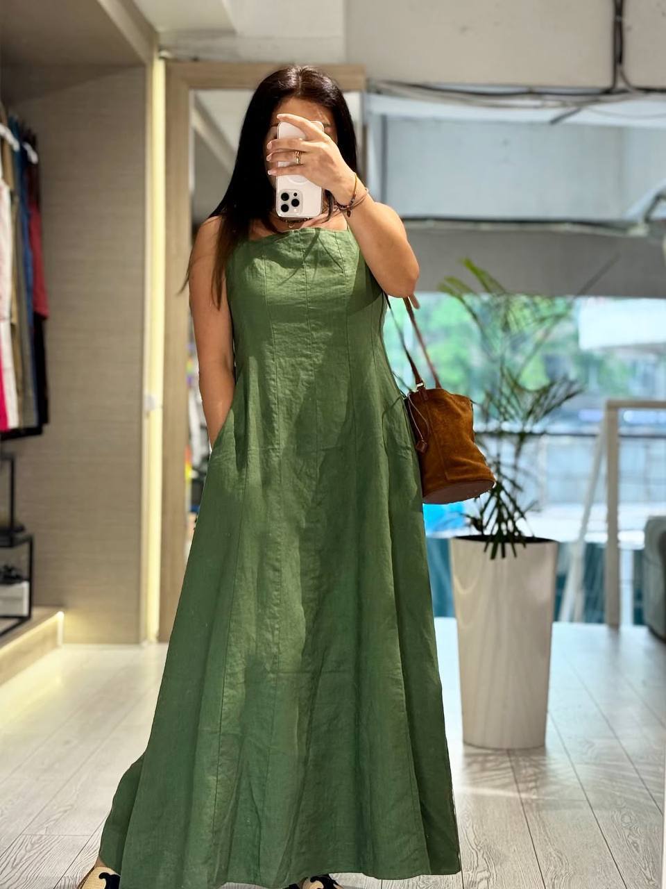 A woman takes a mirror selfie in a boutique wearing a sleeveless, floor-length sage green dress with pockets. She is holding a brown leather shoulder bag and wearing black sandals. The background shows a clothing rack on the left and a potted plant on the right.