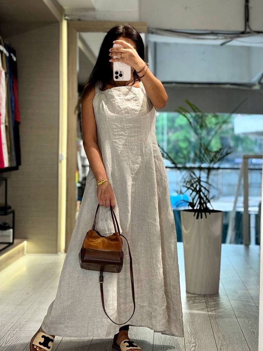 A woman takes a mirror selfie in a clothing store while wearing a light beige linen maxi dress and carrying a brown leather handbag.