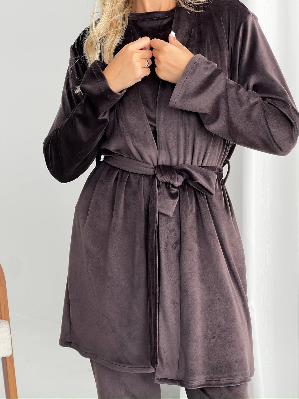 A woman wearing a dark plum velvet robe with a tie belt, shown from the shoulders down against a light gray indoor background.