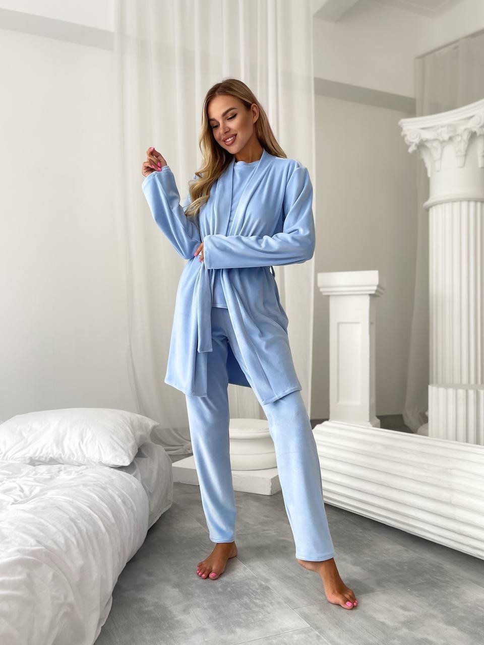 A woman modeling a light blue two-piece pajama set consisting of a long-sleeve robe and matching pants, standing in a bright bedroom with white bedding and classical-style columns.