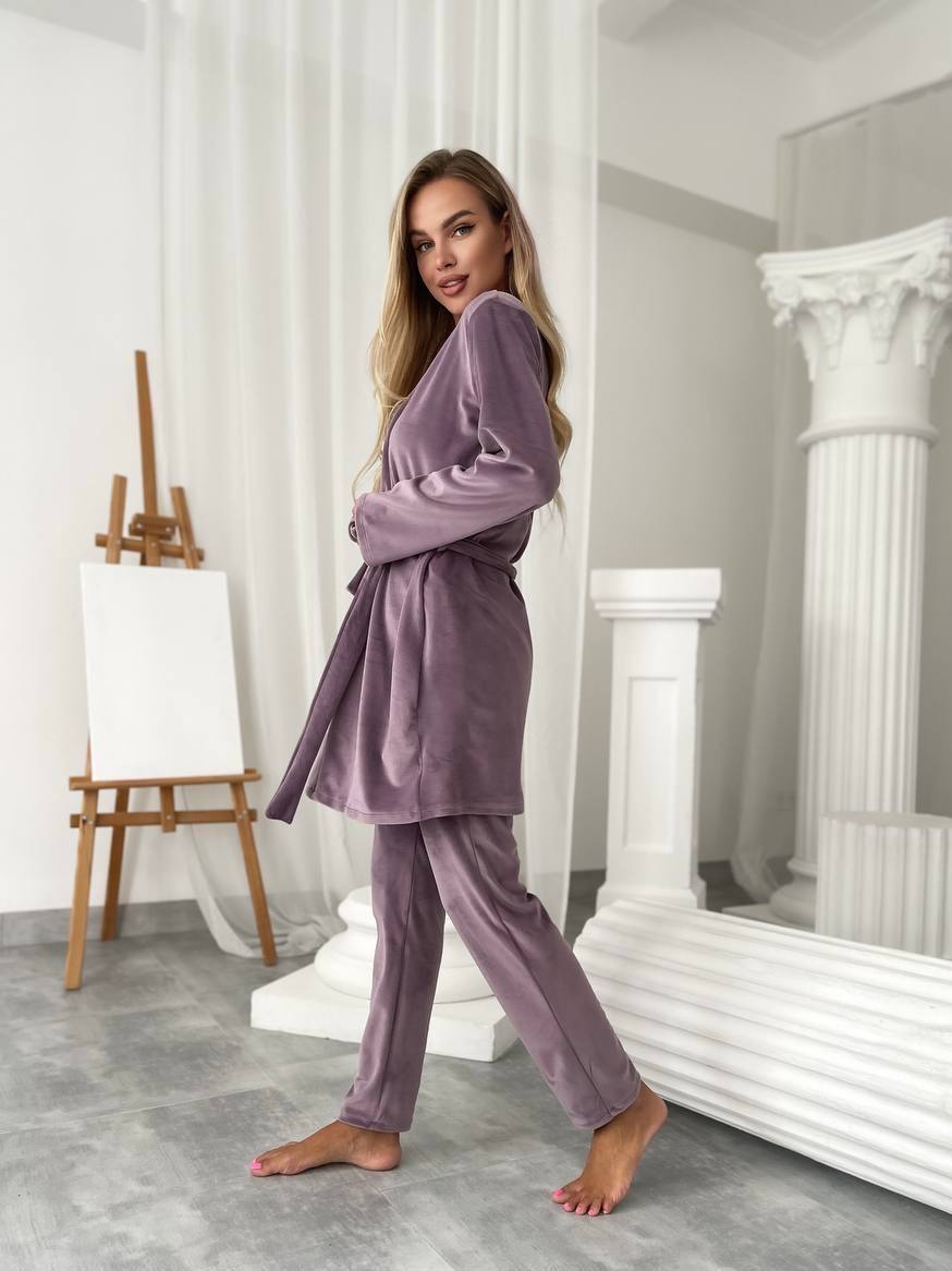 A woman modeling a matching two-piece pajama set in a bright, modern studio with white classical-style columns and an easel. The set consists of a long-sleeve belted robe and matching pants in a dusty lavender-purple velvet-like fabric.